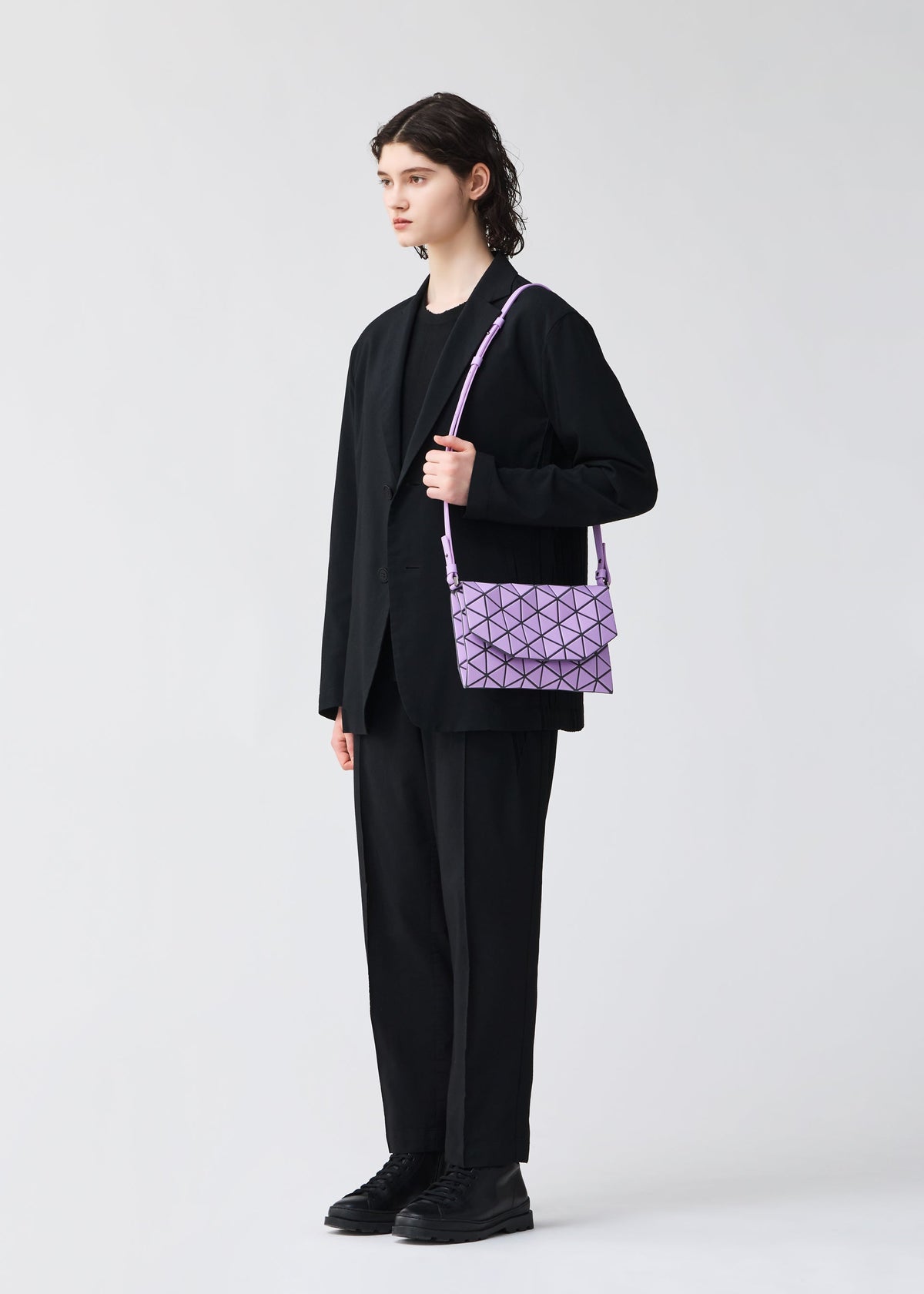 A person with short, dark, wavy hair stands in a black suit and shoes, holding the BAO BAO ISSEY MIYAKE TONNEAU MATTE purple geometric-patterned shoulder bag against a plain light background. view 8 of 8