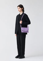 A person with short, dark, wavy hair stands in a black suit and shoes, holding the BAO BAO ISSEY MIYAKE TONNEAU MATTE purple geometric-patterned shoulder bag against a plain light background.