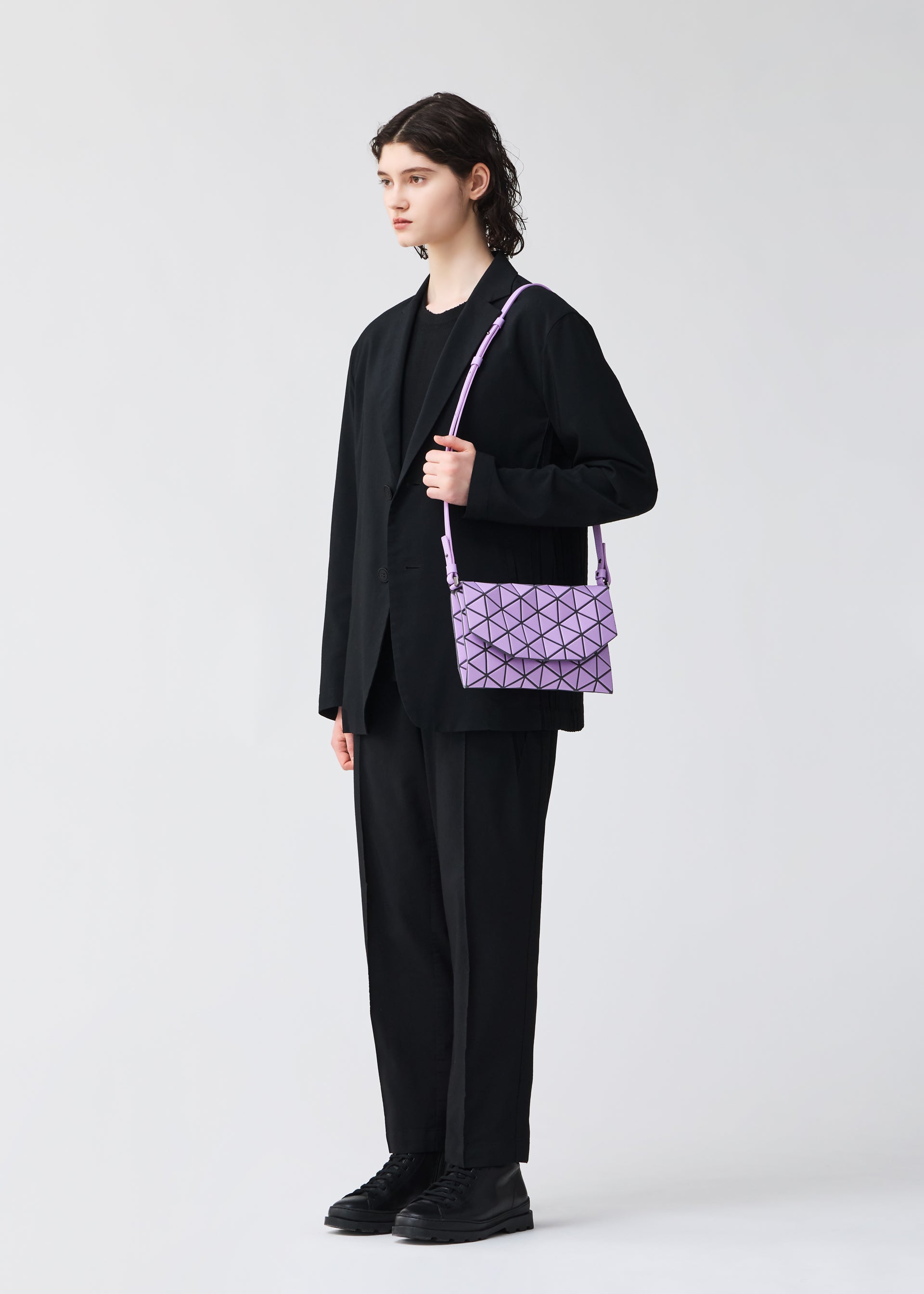 Zoomed view A person with short, dark, wavy hair stands in a black suit and shoes, holding the BAO BAO ISSEY MIYAKE TONNEAU MATTE purple geometric-patterned shoulder bag against a plain light background.