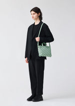 A person with shoulder-length hair in a black suit, shirt, and shoes stands against a white background, carrying the BAO BAO ISSEY MIYAKE TONNEAU MATTE—a small, barrel-shaped mini handbag with a mint green geometric pattern—over one shoulder.