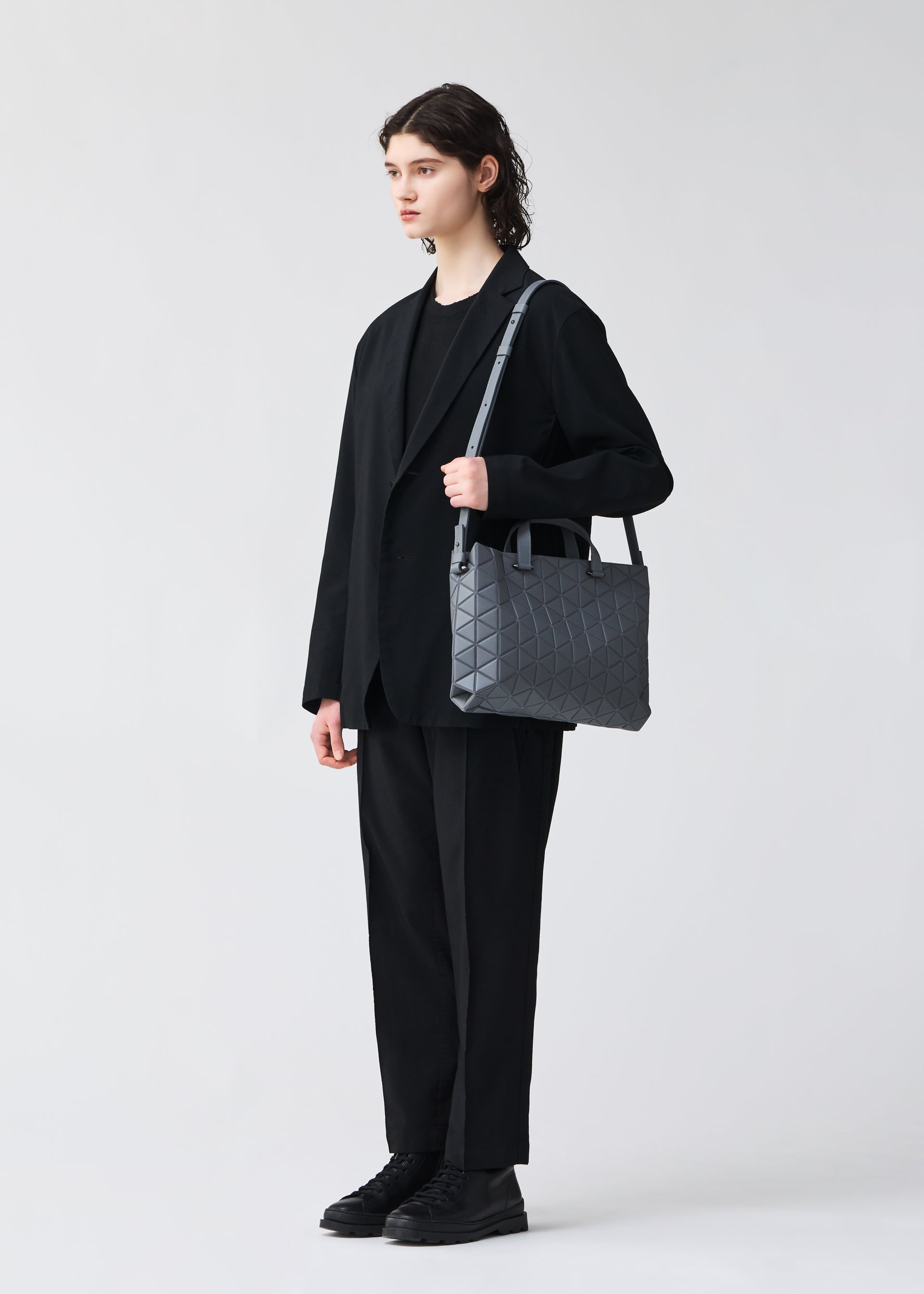 Zoomed view A person with shoulder-length dark hair stands against a plain background, dressed in a loose black suit and shoes, carrying the BAO BAO ISSEY MIYAKE TONNEAU MATTE A4 barrel-shaped geometric handbag over their shoulder.