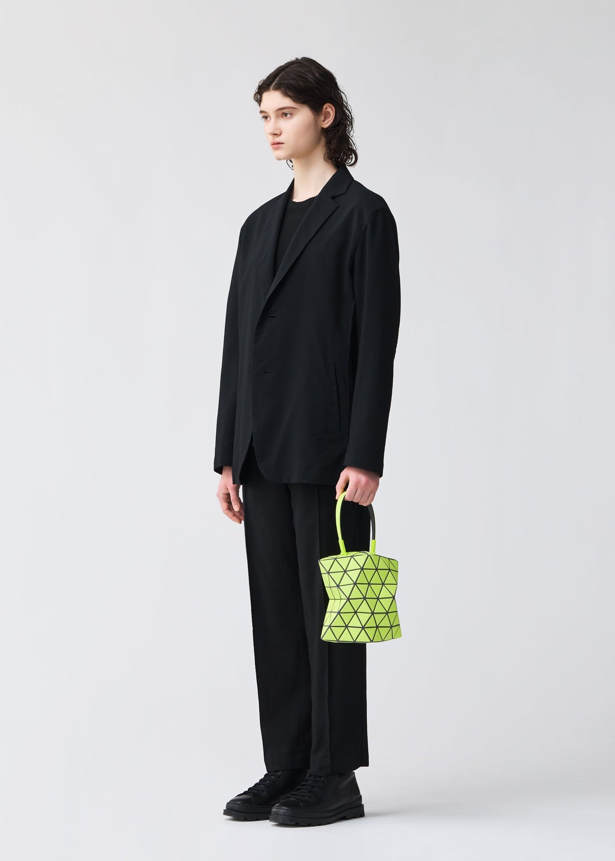 A person with shoulder-length hair wears a loose black suit and shoes, standing against a plain light background, holding the BAO BAO ISSEY MIYAKE TORSO—a small, neon yellow equilateral triangle handbag with a geometric pattern. view 8 of 8
