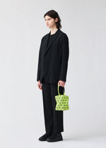 A person with shoulder-length hair wears a loose black suit and shoes, standing against a plain light background, holding the BAO BAO ISSEY MIYAKE TORSO—a small, neon yellow equilateral triangle handbag with a geometric pattern.