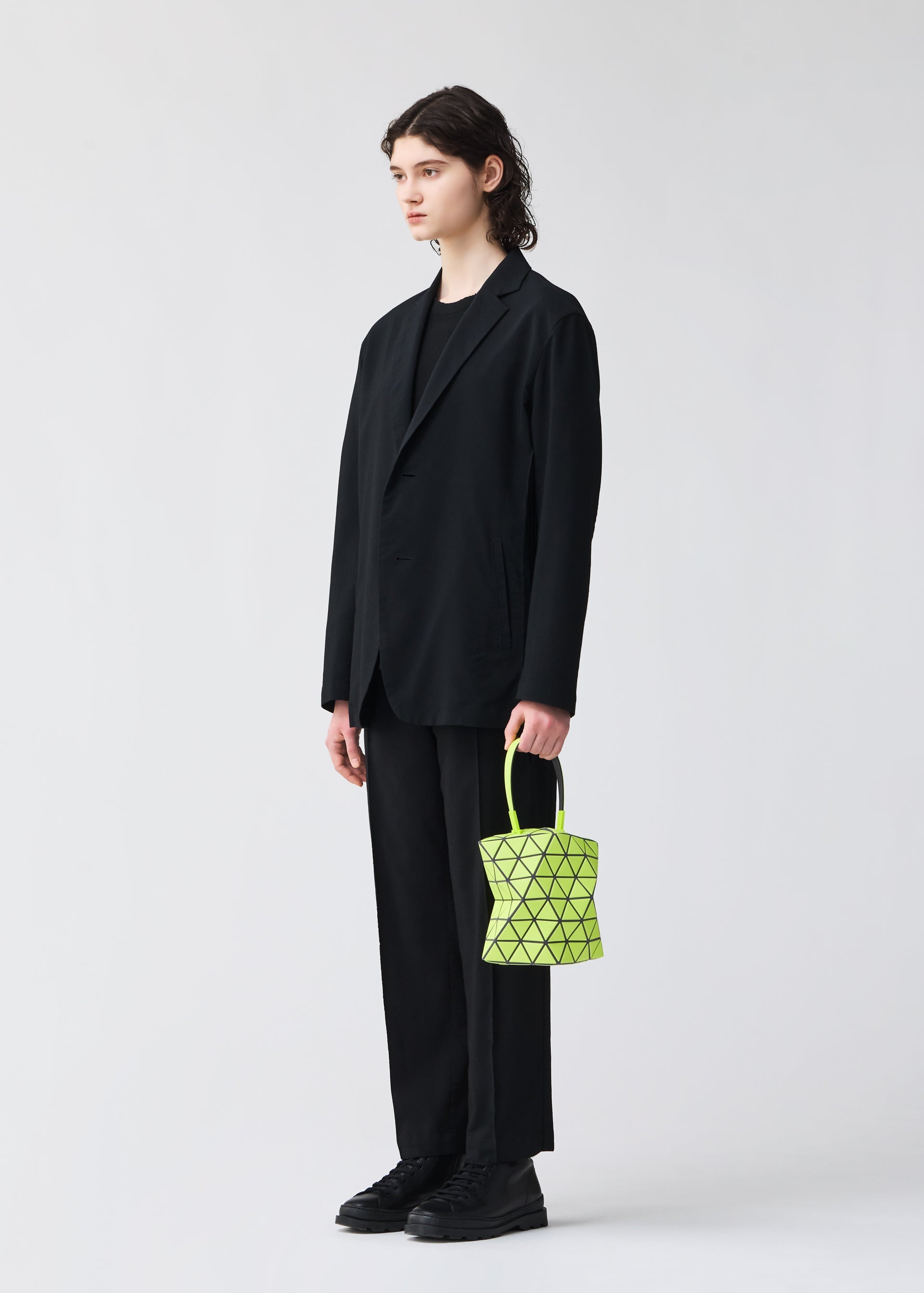 Zoomed view A person with shoulder-length hair wears a loose black suit and shoes, standing against a plain light background, holding the BAO BAO ISSEY MIYAKE TORSO—a small, neon yellow equilateral triangle handbag with a geometric pattern.