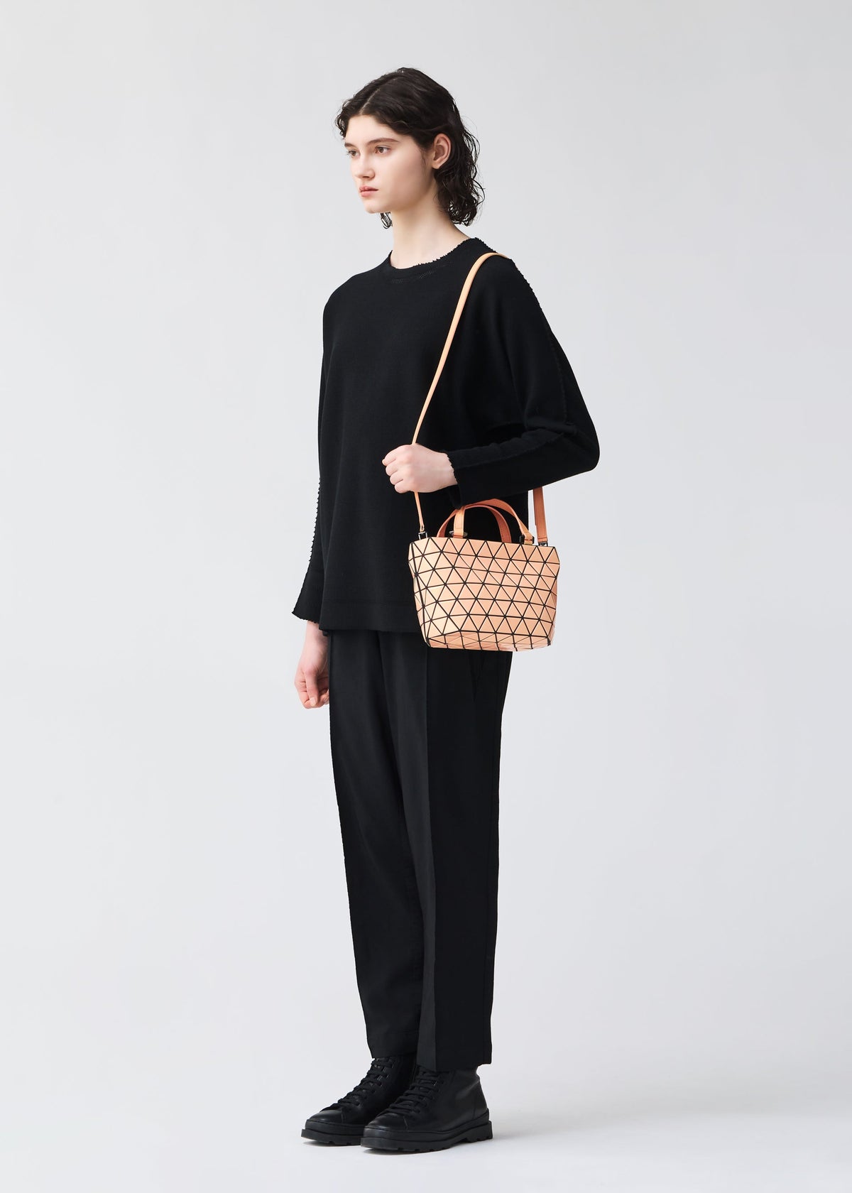 Against a light gray background, a person with short, dark hair holds the BAO BAO ISSEY MIYAKE CRYSTAL GLOSS mini handbag, styled with a loose black sweater, black pants, and boots. view 6 of 6