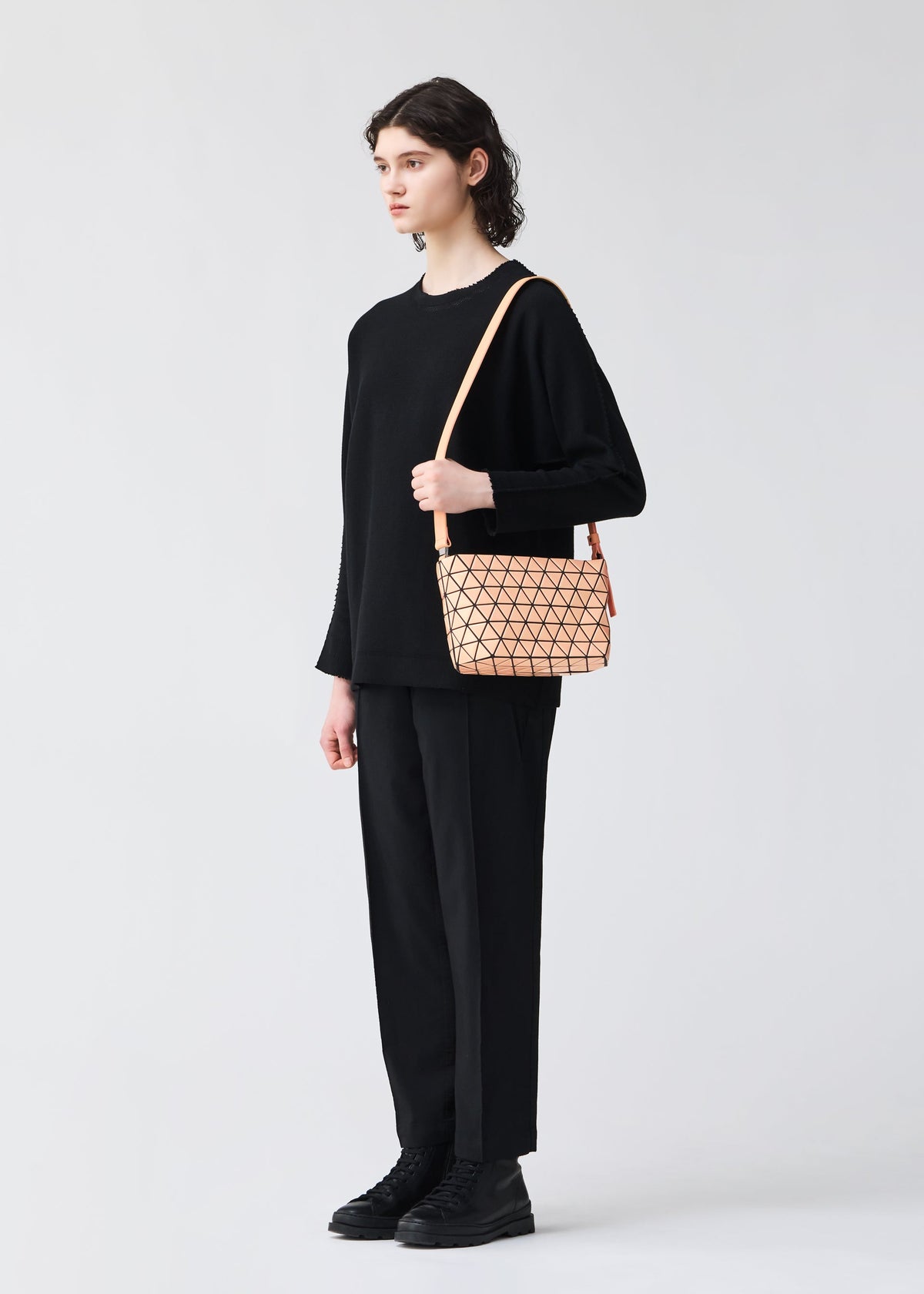 A person with short, dark hair stands against a plain background, dressed in all black and carrying the BAO BAO ISSEY MIYAKE CRYSTAL GLOSS—a small beige geometric shoulder bag with a leather strap. view 6 of 6