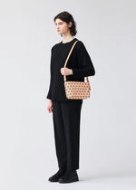 A person with short, dark hair stands against a plain background, dressed in all black and carrying the BAO BAO ISSEY MIYAKE CRYSTAL GLOSS—a small beige geometric shoulder bag with a leather strap.