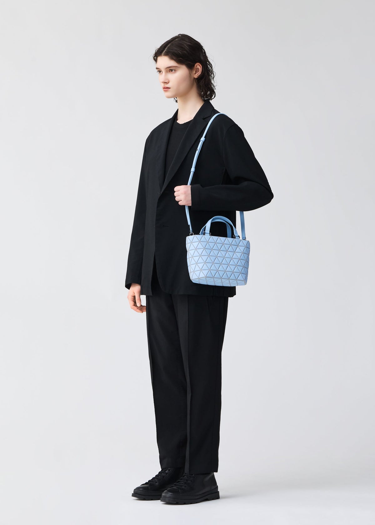A person with shoulder-length dark hair in a black suit and shoes holds the BAO BAO ISSEY MIYAKE CRYSTAL MATTE light blue mini handbag with a geometric pattern and 2-way leather straps, standing against a plain light background. view 7 of 7