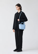 A person dressed in a black suit and black shoes stands against a plain light background, holding the BAO BAO ISSEY MIYAKE CRYSTAL MATTE mini handbag—a small, light blue bag featuring geometric patterns, leather straps, and a shoulder strap.