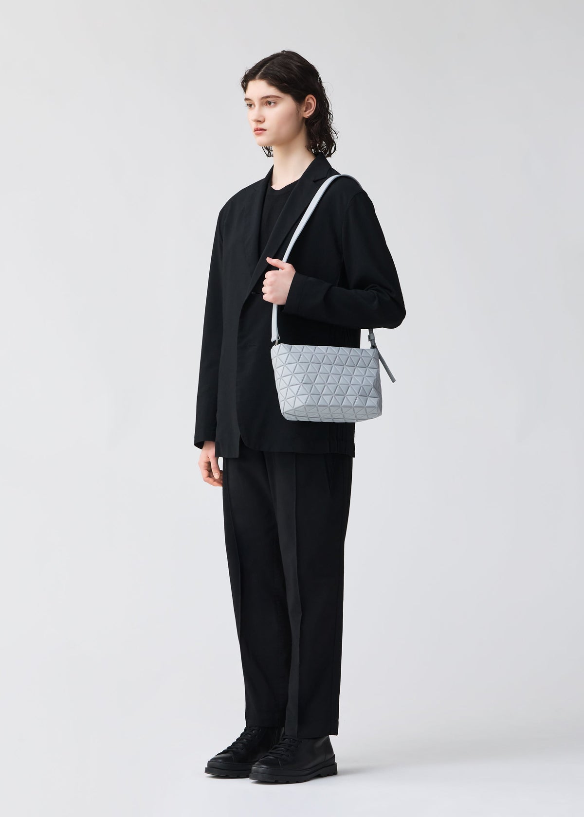 A person with medium-length brown hair in a loose black suit and shoes stands against a light background, carrying the BAO BAO ISSEY MIYAKE CRYSTAL MATTE light gray quilted shoulder bag with leather straps. view 7 of 7