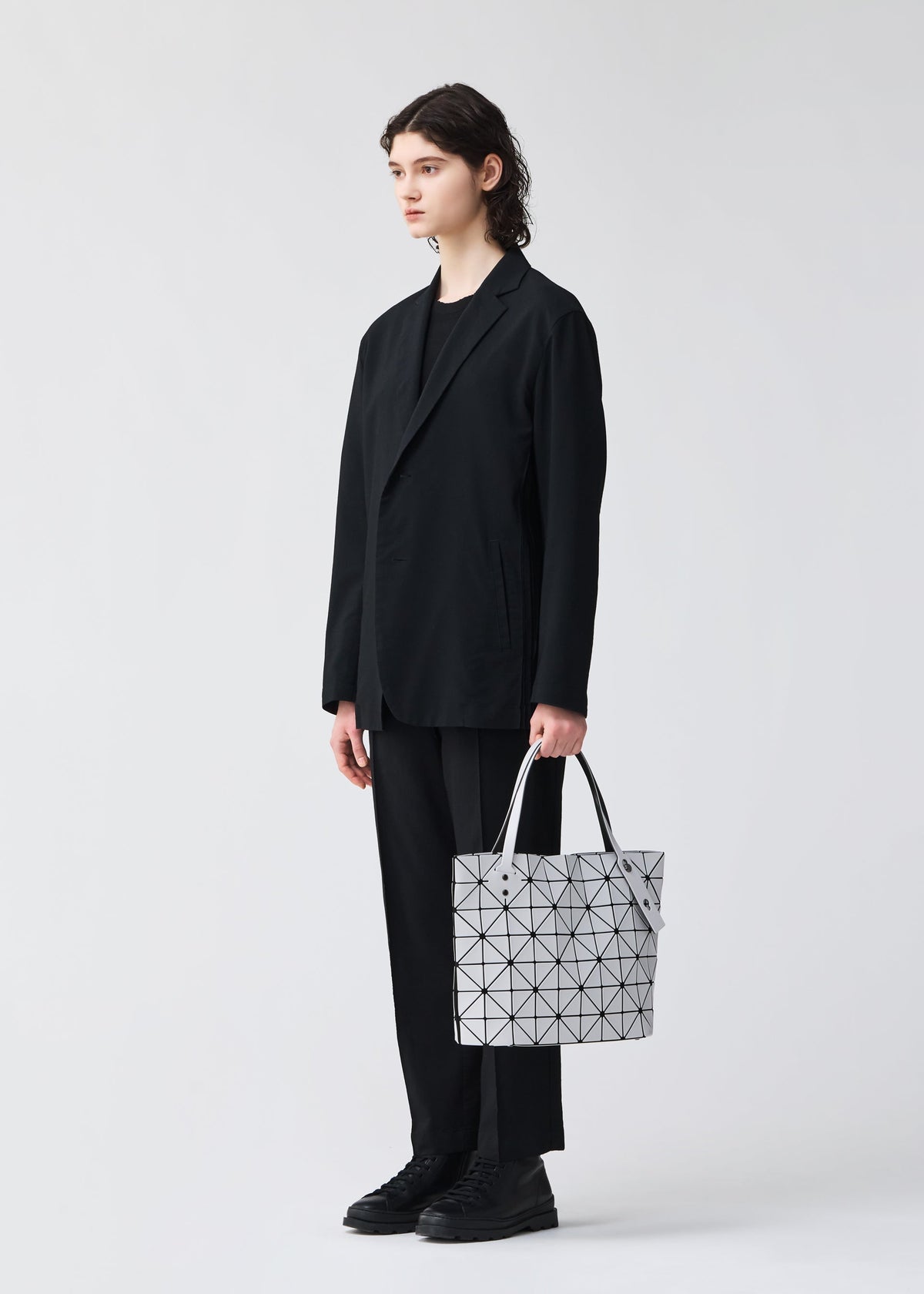 A person with shoulder-length hair, in a loose black suit and black shoes, faces left holding the ROCK MATTE bag by BAO BAO ISSEY MIYAKE—its white geometric design fits a 13-inch laptop—against a plain light background. view 7 of 8