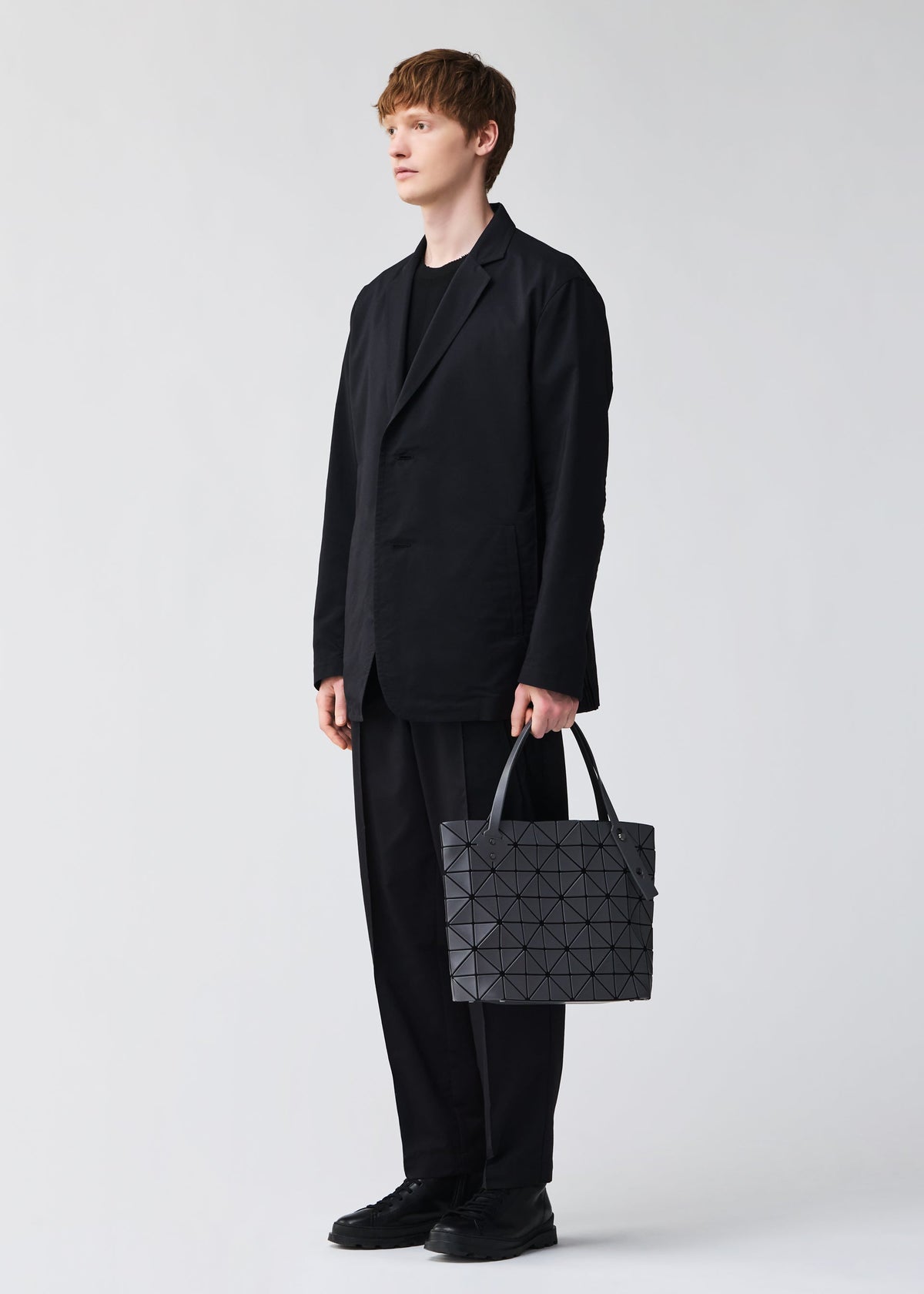 Dressed in a loose black suit and shoes, a person holds the BAO BAO ISSEY MIYAKE ROCK MATTE handbag—its geometric gray design and gusseted bottom fit a 13-inch laptop—posing against a plain light background. view 8 of 8