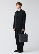 Dressed in a loose black suit and shoes, a person holds the BAO BAO ISSEY MIYAKE ROCK MATTE handbag—its geometric gray design and gusseted bottom fit a 13-inch laptop—posing against a plain light background.