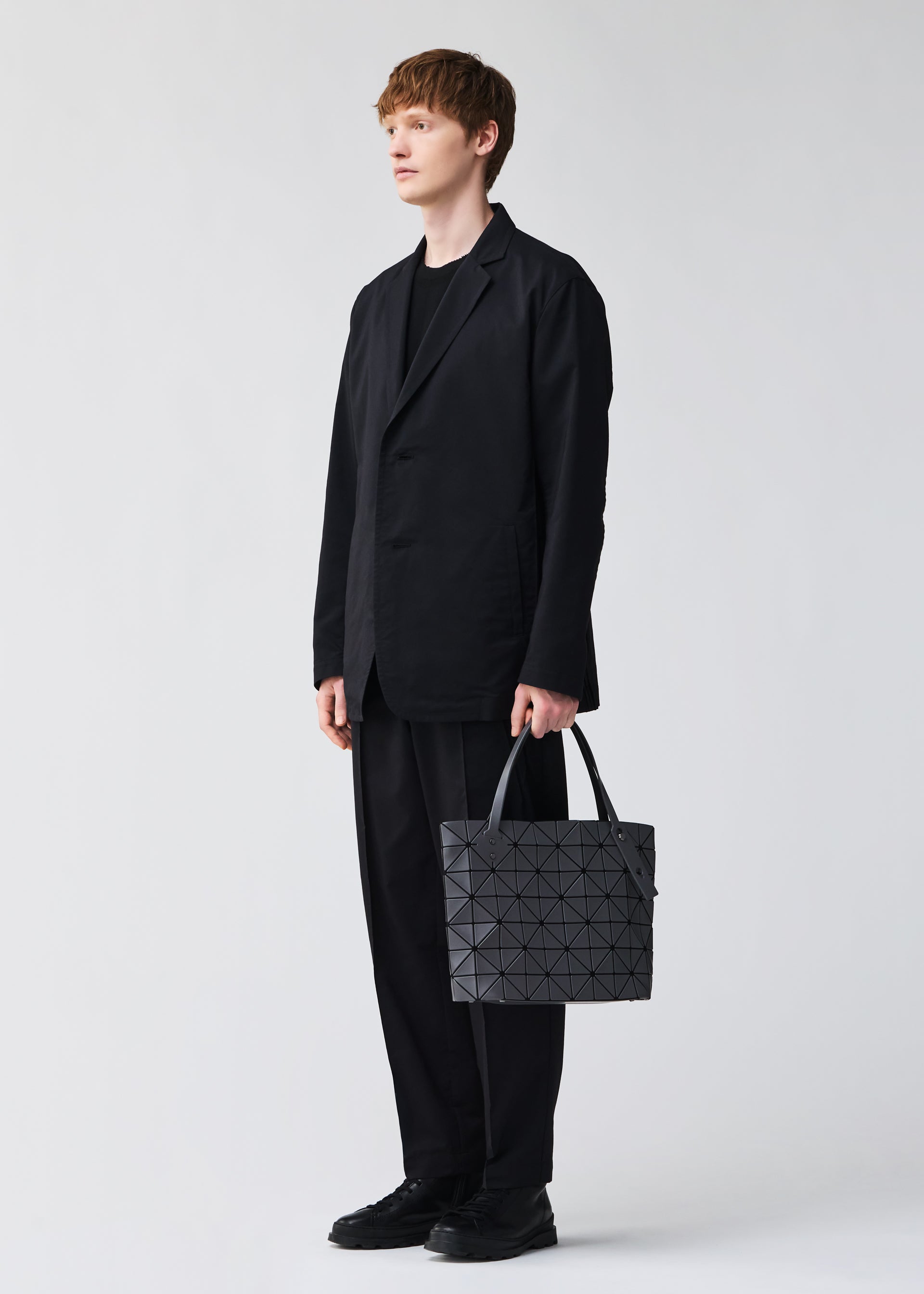 Zoomed view Dressed in a loose black suit and shoes, a person holds the BAO BAO ISSEY MIYAKE ROCK MATTE handbag—its geometric gray design and gusseted bottom fit a 13-inch laptop—posing against a plain light background.