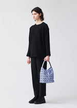 A person with short, dark hair stands against a plain light background, dressed in all black and holding the BAO BAO ISSEY MIYAKE VALLEY handbag—a geometric-patterned bag in a light purple hue—in their left hand.