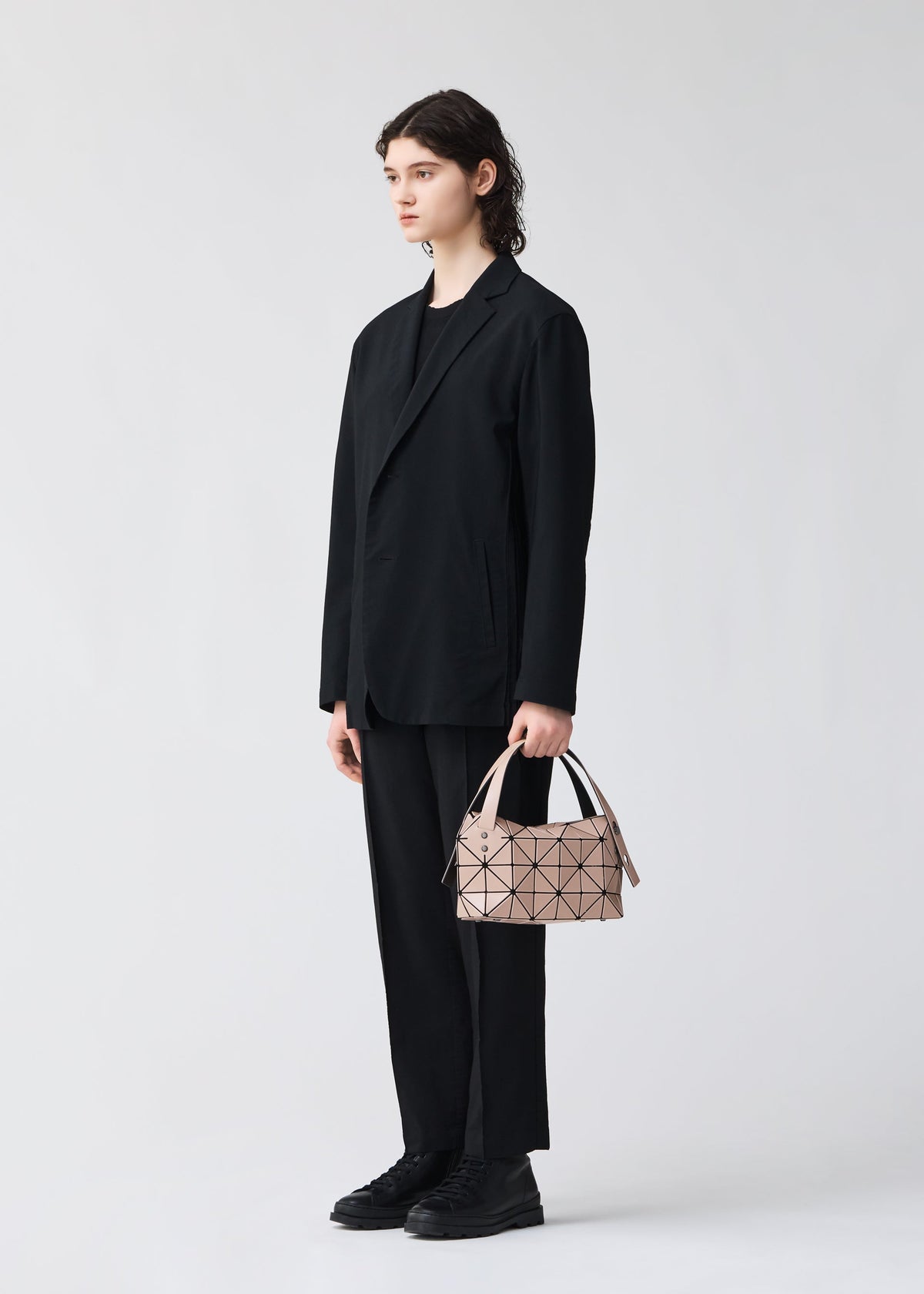 A person with shoulder-length dark hair, wearing a loose black suit and shoes, stands against a light background holding the BAO BAO ISSEY MIYAKE BOSTON mini bag featuring modern geometric patterns in their left hand. view 6 of 6