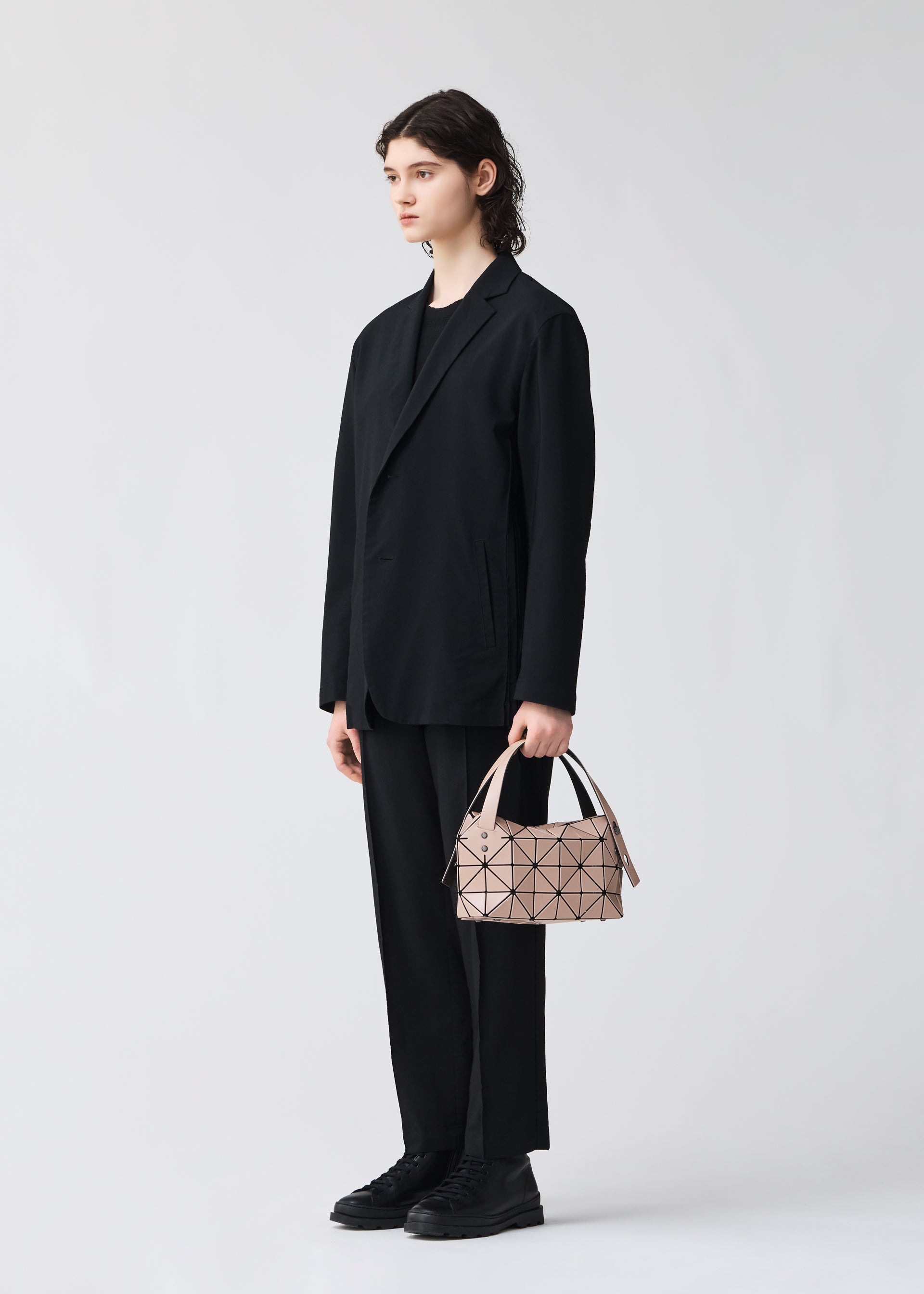 Zoomed view A person with shoulder-length dark hair, wearing a loose black suit and shoes, stands against a light background holding the BAO BAO ISSEY MIYAKE BOSTON mini bag featuring modern geometric patterns in their left hand.