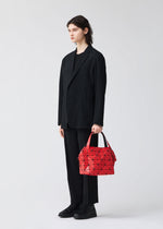 A person with long dark hair in a loose black suit stands against a plain light background, holding the geometric red BOSTON bag from BAO BAO ISSEY MIYAKE in their left hand—a striking example of modern design and spacious storage.