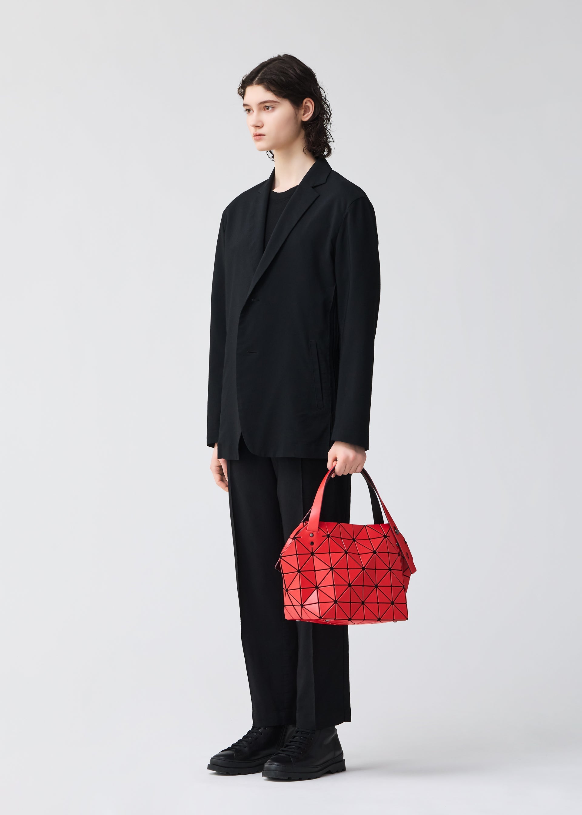 Zoomed view A person with long dark hair in a loose black suit stands against a plain light background, holding the geometric red BOSTON bag from BAO BAO ISSEY MIYAKE in their left hand—a striking example of modern design and spacious storage.