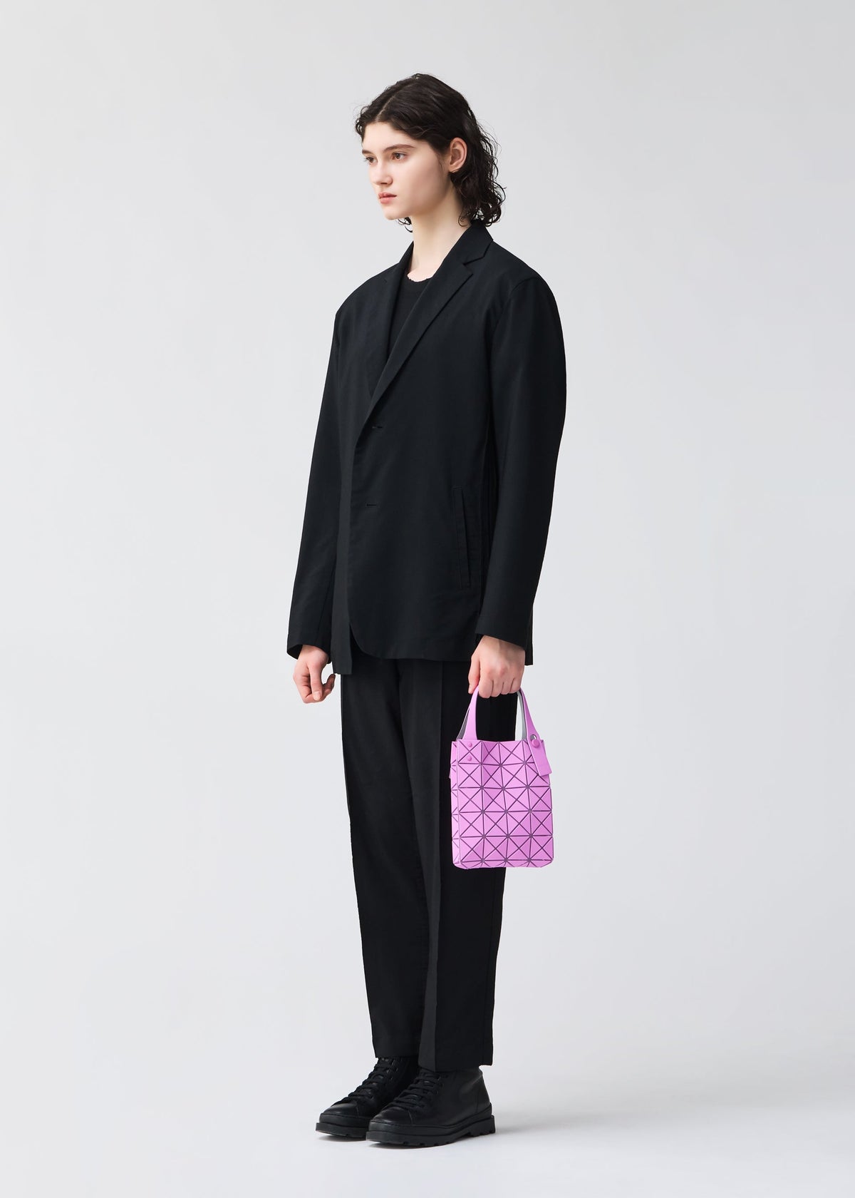 A person with long dark hair, wearing a loose black suit and black shoes, stands against a light background holding the BAO BAO ISSEY MIYAKE PRISM PLUS mini tote bag in pink with a geometric design. view 9 of 10