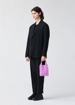 A person with long dark hair, wearing a loose black suit and black shoes, stands against a light background holding the BAO BAO ISSEY MIYAKE PRISM PLUS mini tote bag in pink with a geometric design.