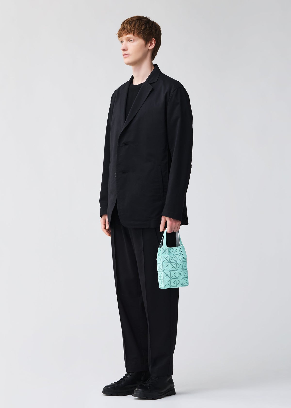 A person with short light brown hair wears a loose black suit and shoes, standing against a plain light background, holding the compact PRISM PLUS tote from BAO BAO ISSEY MIYAKE—its small light blue geometric pattern adds a bold accent. view 10 of 10