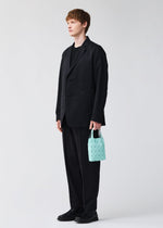 A person with short light brown hair wears a loose black suit and shoes, standing against a plain light background, holding the compact PRISM PLUS tote from BAO BAO ISSEY MIYAKE—its small light blue geometric pattern adds a bold accent.