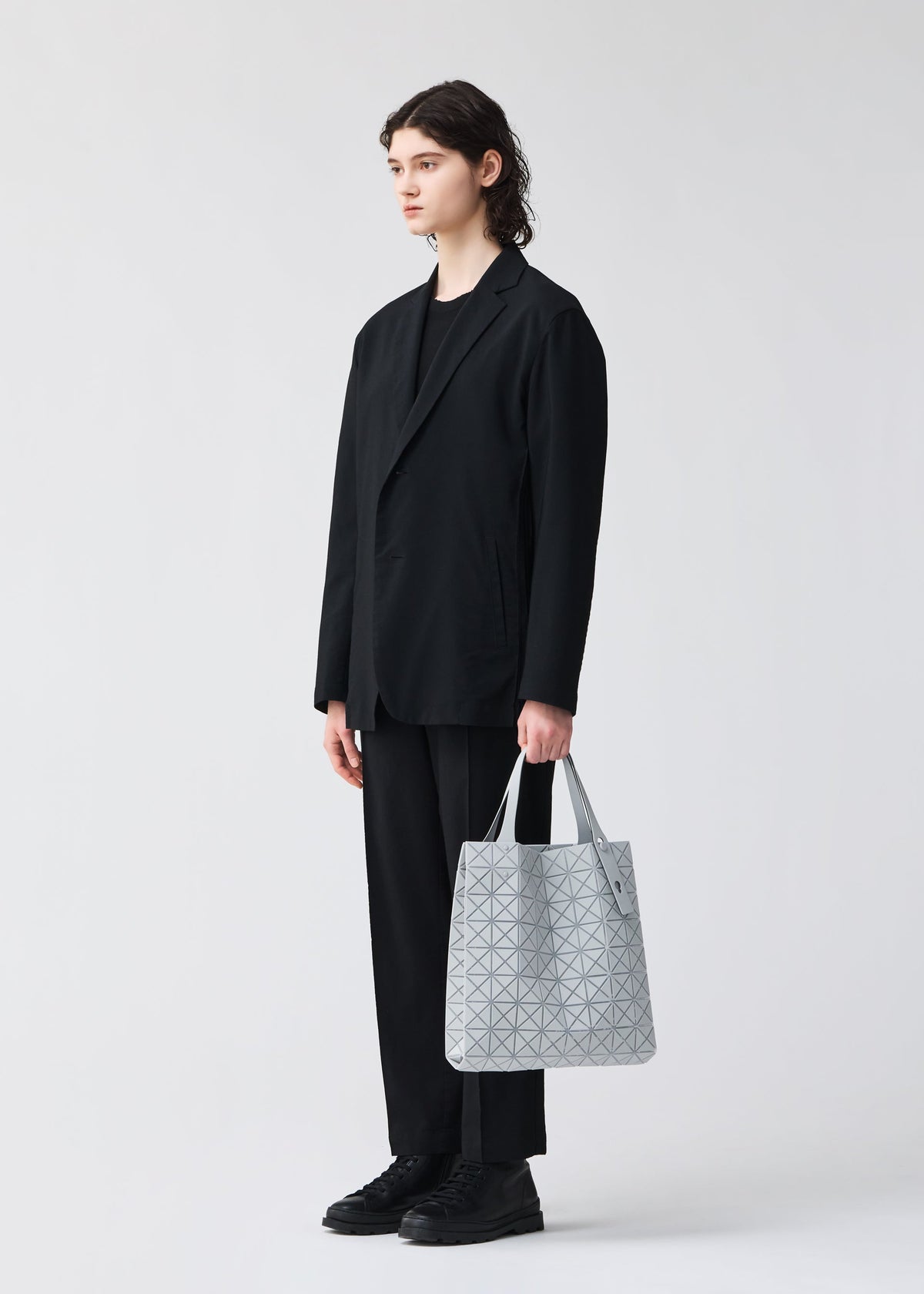 A person with long brown hair, in a loose black suit and black shoes, stands holding the light gray PRISM PLUS tote by BAO BAO ISSEY MIYAKE, which features a geometric pattern and subtle fabric grain against a plain light background. view 9 of 10