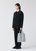 A person with long brown hair, in a loose black suit and black shoes, stands holding the light gray PRISM PLUS tote by BAO BAO ISSEY MIYAKE, which features a geometric pattern and subtle fabric grain against a plain light background.