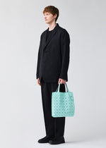 A person with short light brown hair, dressed in a loose black suit and shoes, stands against a plain background holding the BAO BAO ISSEY MIYAKE PRISM PLUS tote bag with a light blue geometric pattern and notable storage in their left hand.