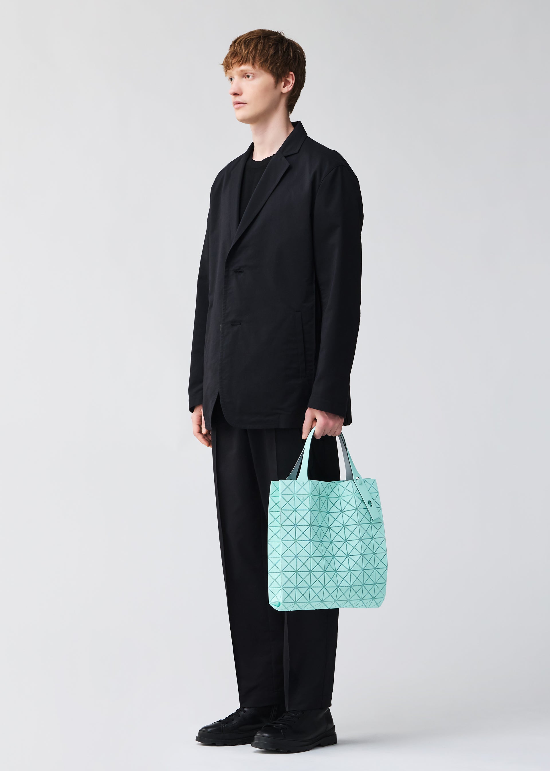 Zoomed view A person with short light brown hair, dressed in a loose black suit and shoes, stands against a plain background holding the BAO BAO ISSEY MIYAKE PRISM PLUS tote bag with a light blue geometric pattern and notable storage in their left hand.