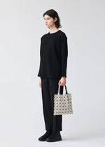A person with short dark hair, dressed in a loose black sweater, black pants, and black shoes, holds the BAO BAO ISSEY MIYAKE PRISM FROST tote bag with a geometric pattern against a plain light background.