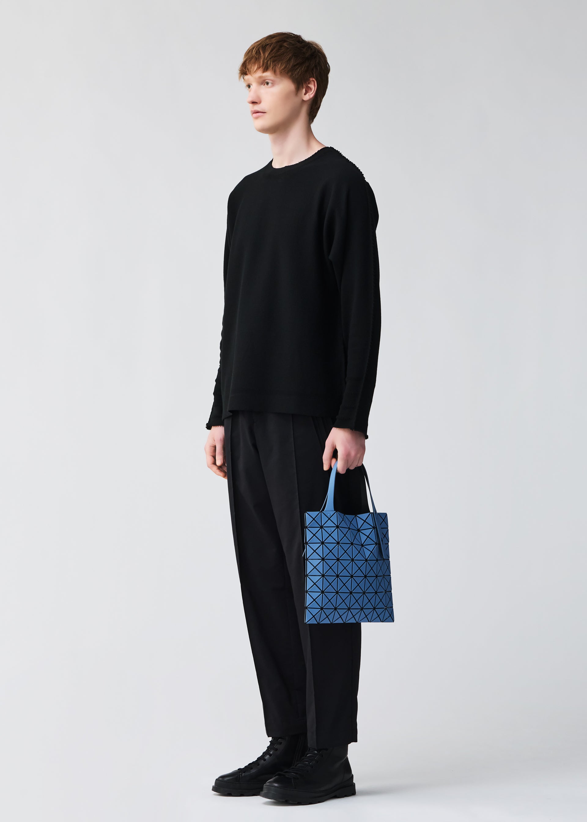 Zoomed view A person with short light brown hair, dressed in black long sleeves, loose black pants, and black shoes, stands facing left holding a blue PRISM FROST tote bag by BAO BAO ISSEY MIYAKE. The background is light gray.