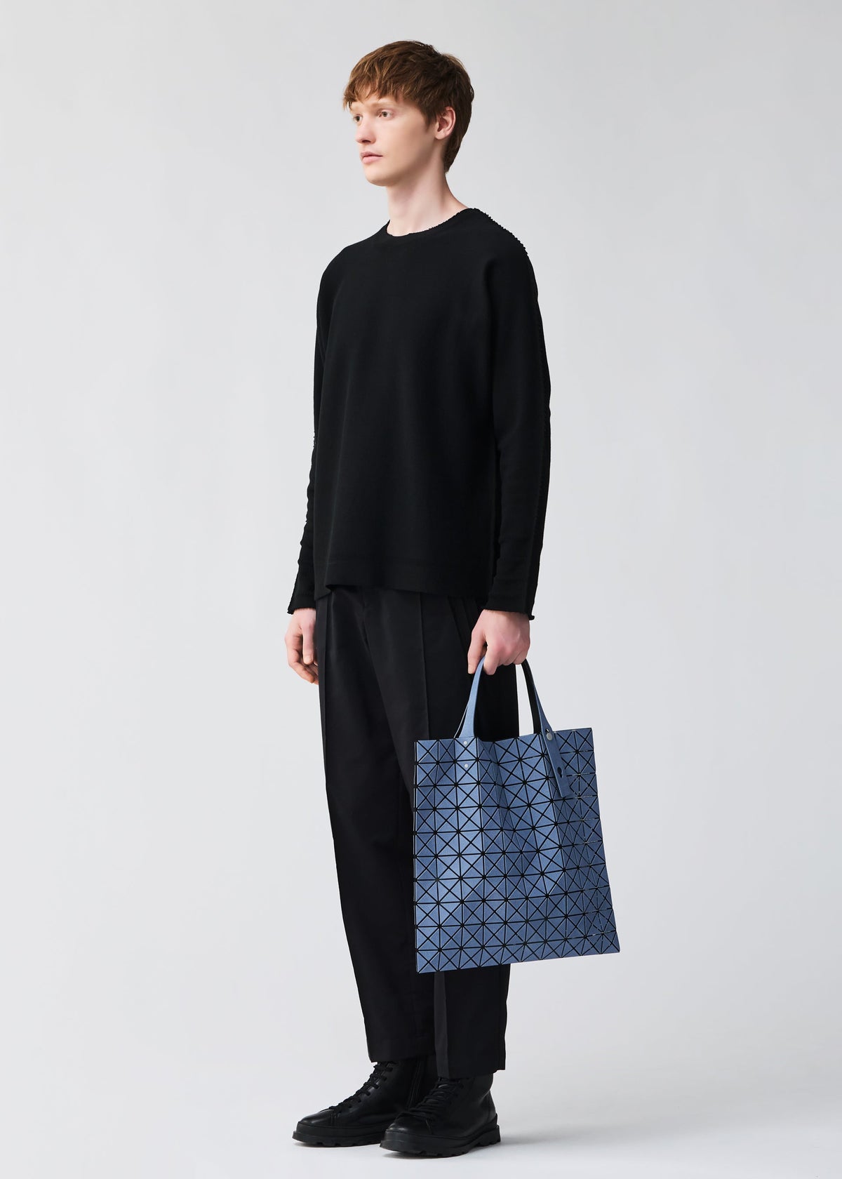 A person with short, light brown hair stands against a plain light background, wearing black attire and holding the BAO BAO ISSEY MIYAKE PRISM DENIM tote bag in blue geometric-patterned denim with a zippered interior pocket. view 7 of 7