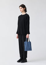 A person with short dark hair, dressed in a loose black sweater, black pants, and black shoes, stands against a plain light background holding a small blue PRISM DENIM tote bag by BAO BAO ISSEY MIYAKE.