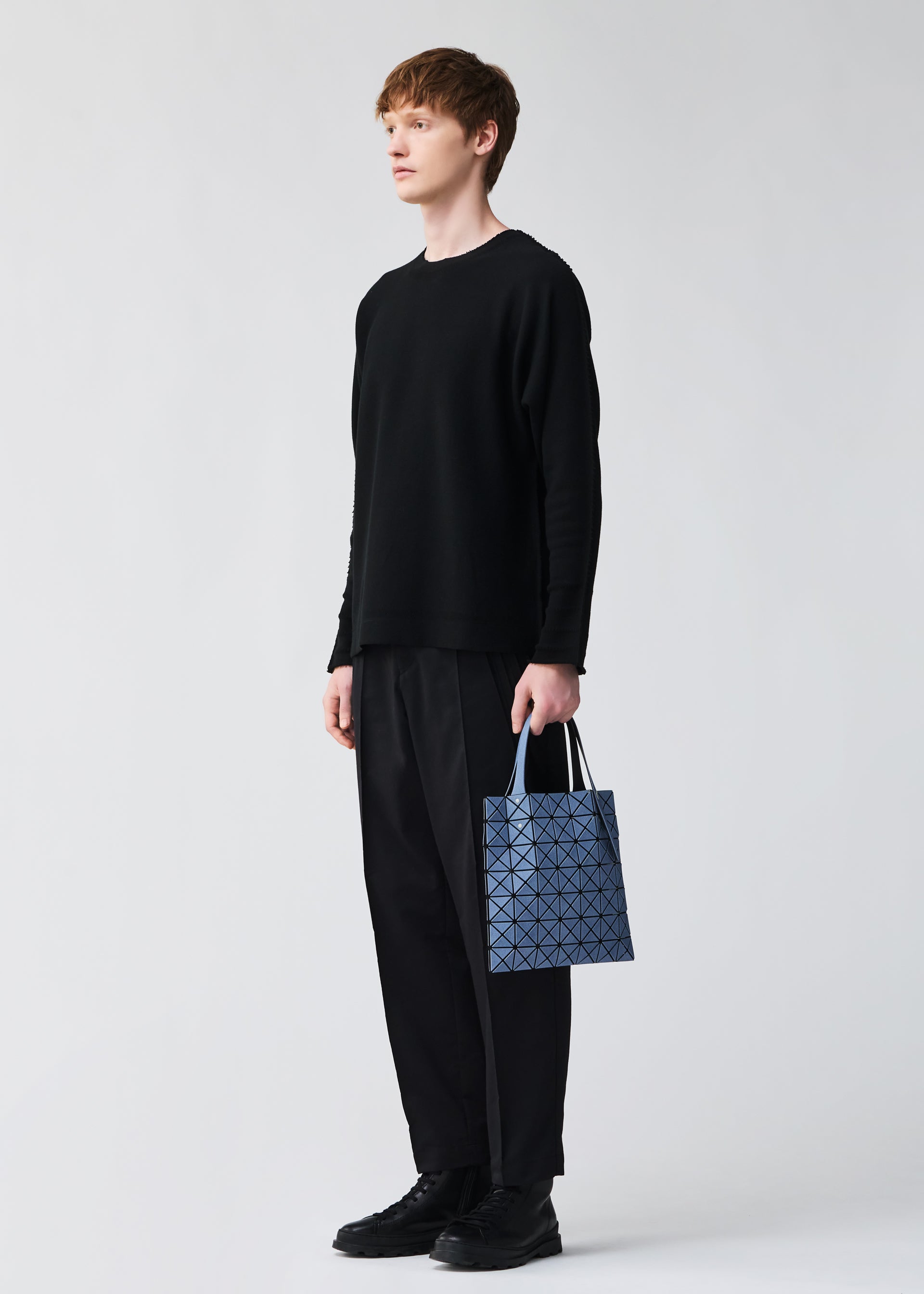 Zoomed view A person in black long-sleeve top and pants holds a small blue BAO BAO ISSEY MIYAKE PRISM DENIM tote bag with a zippered inside pocket, set against a plain light gray background.