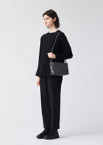 A person with short, dark hair wears a loose black sweater, black pants, and black shoes, holding the BAO BAO ISSEY MIYAKE LOOP ONE-TONE shoulder bag with adjustable strap and geometric pattern against a plain light background.