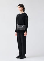 A person with short dark hair stands against a plain background, wearing black clothes and platform shoes, carrying the BAO BAO ISSEY MIYAKE LOOP ONE-TONE geometric-patterned shoulder bag with an adjustable strap.