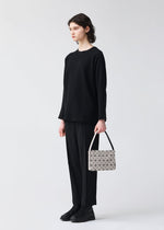 A person with short, dark hair stands against a plain background, dressed in black attire and holding the OBLONG handbag by BAO BAO ISSEY MIYAKE—a small white minimalist bag featuring a geometric pattern.