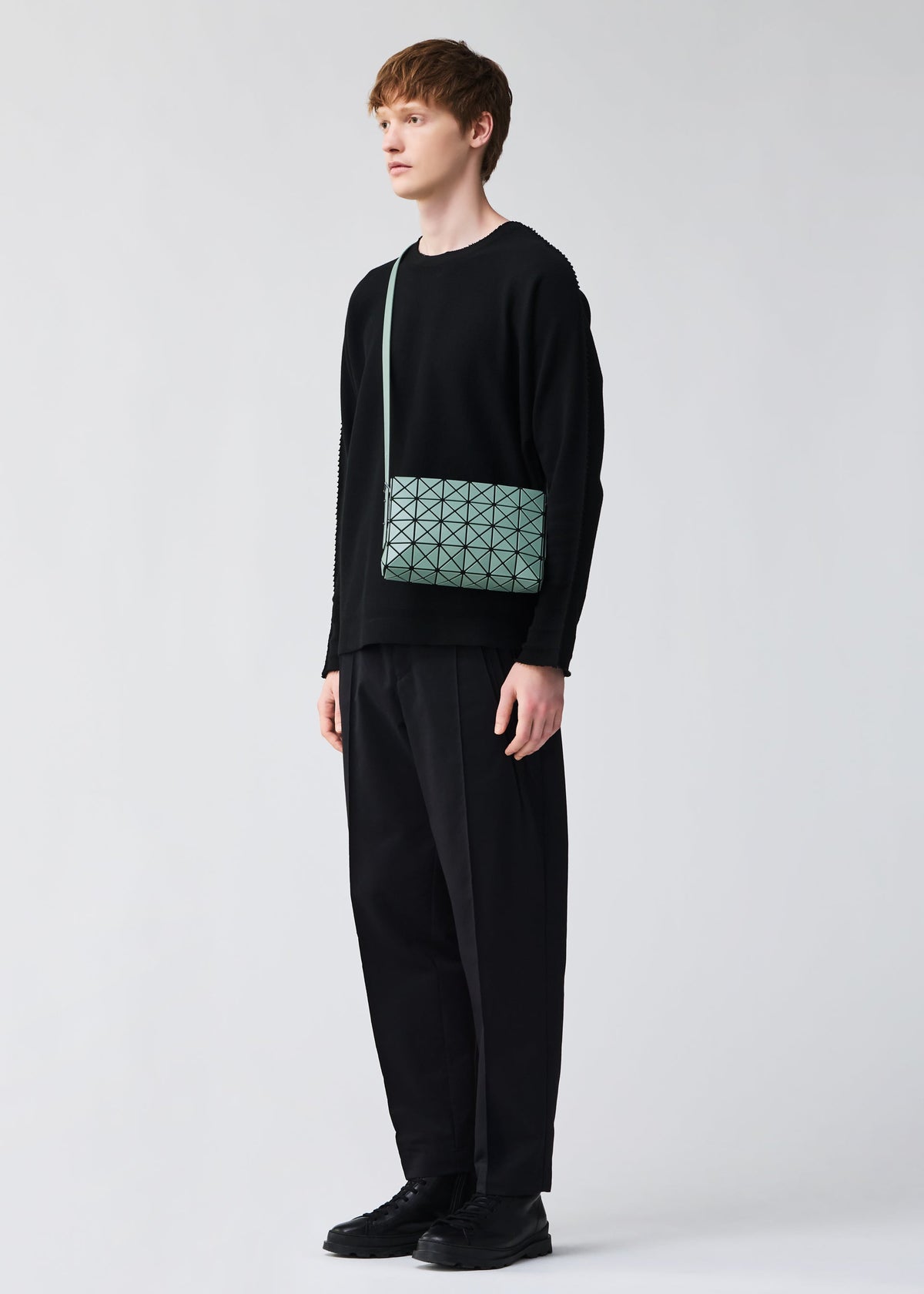 A young person stands against a plain light background, wearing a loose black sweater, wide black pants, black shoes, and the BAO BAO ISSEY MIYAKE OBLONG light green geometric minimalist handbag worn cross-body. view 8 of 8