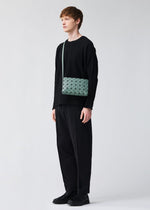 A young person stands against a plain light background, wearing a loose black sweater, wide black pants, black shoes, and the BAO BAO ISSEY MIYAKE OBLONG light green geometric minimalist handbag worn cross-body.