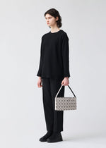 A person stands against a plain light background, dressed in black and holding the OBLONG handbag by BAO BAO ISSEY MIYAKE—a rectangular white bag with a bold black geometric pattern.