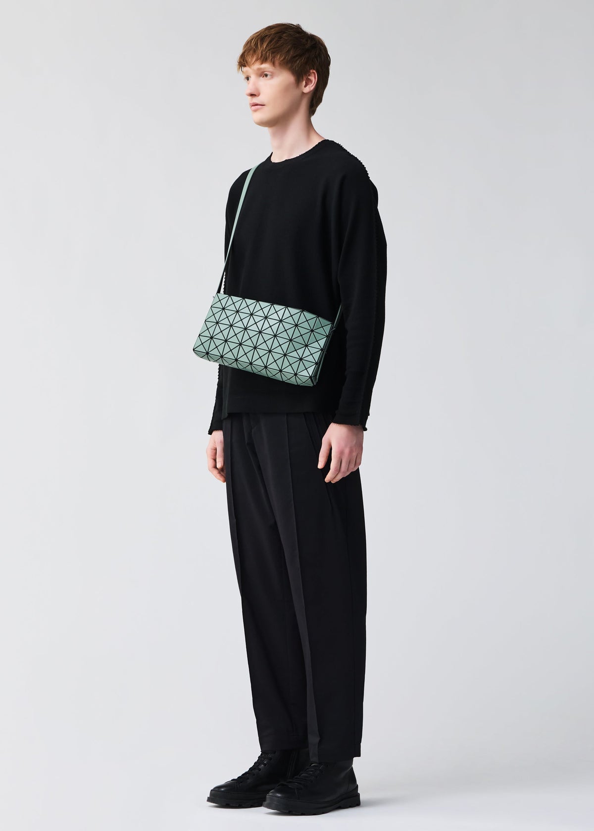 A person with short brown hair in a loose black outfit and black shoes stands against a plain light background, carrying the BAO BAO ISSEY MIYAKE OBLONG geometric-patterned mint green cross-body bag. view 8 of 8