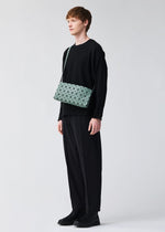 A person with short brown hair in a loose black outfit and black shoes stands against a plain light background, carrying the BAO BAO ISSEY MIYAKE OBLONG geometric-patterned mint green cross-body bag.