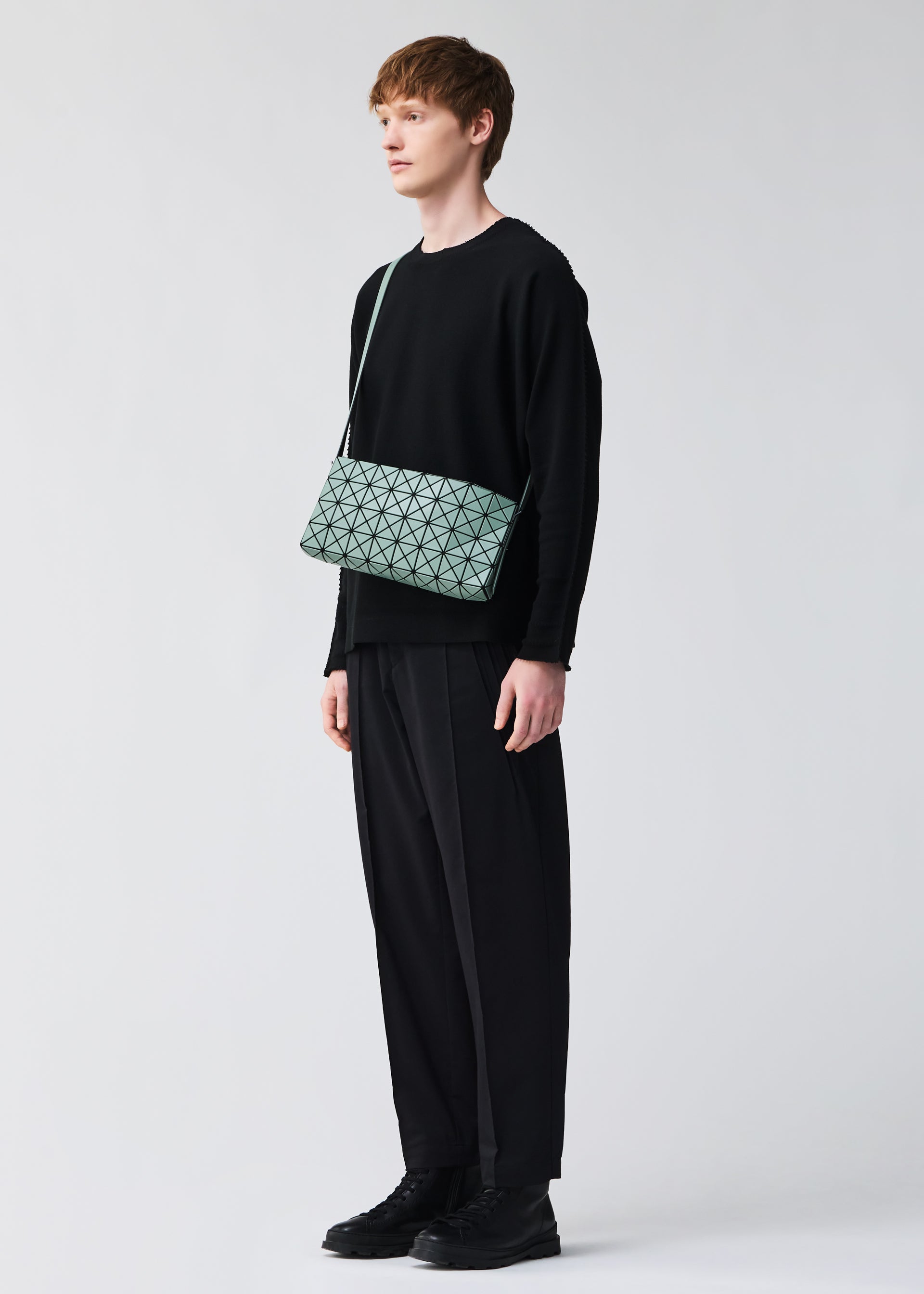 Zoomed view A person with short brown hair in a loose black outfit and black shoes stands against a plain light background, carrying the BAO BAO ISSEY MIYAKE OBLONG geometric-patterned mint green cross-body bag.