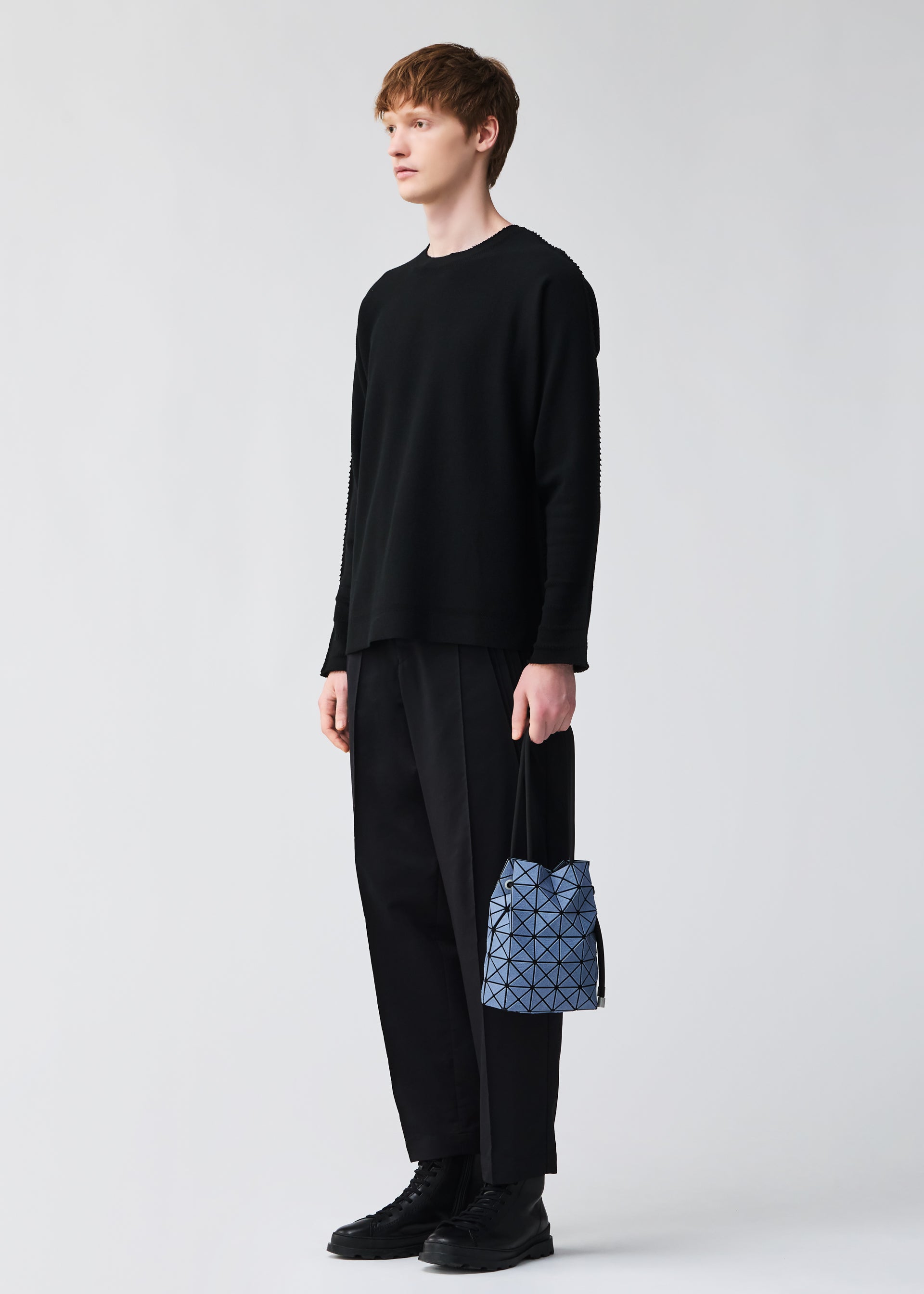 Zoomed view A person holds the BAO BAO ISSEY MIYAKE WRING DENIM, a small geometric-patterned blue and gray drawstring bag, while wearing black attire and standing against a plain light background.