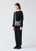 A person with shoulder-length dark hair stands against a plain light background, wearing black clothing and boots, and carrying the LUCENT METALLIC silver geometric shoulder bag by BAO BAO ISSEY MIYAKE with a white strap.
