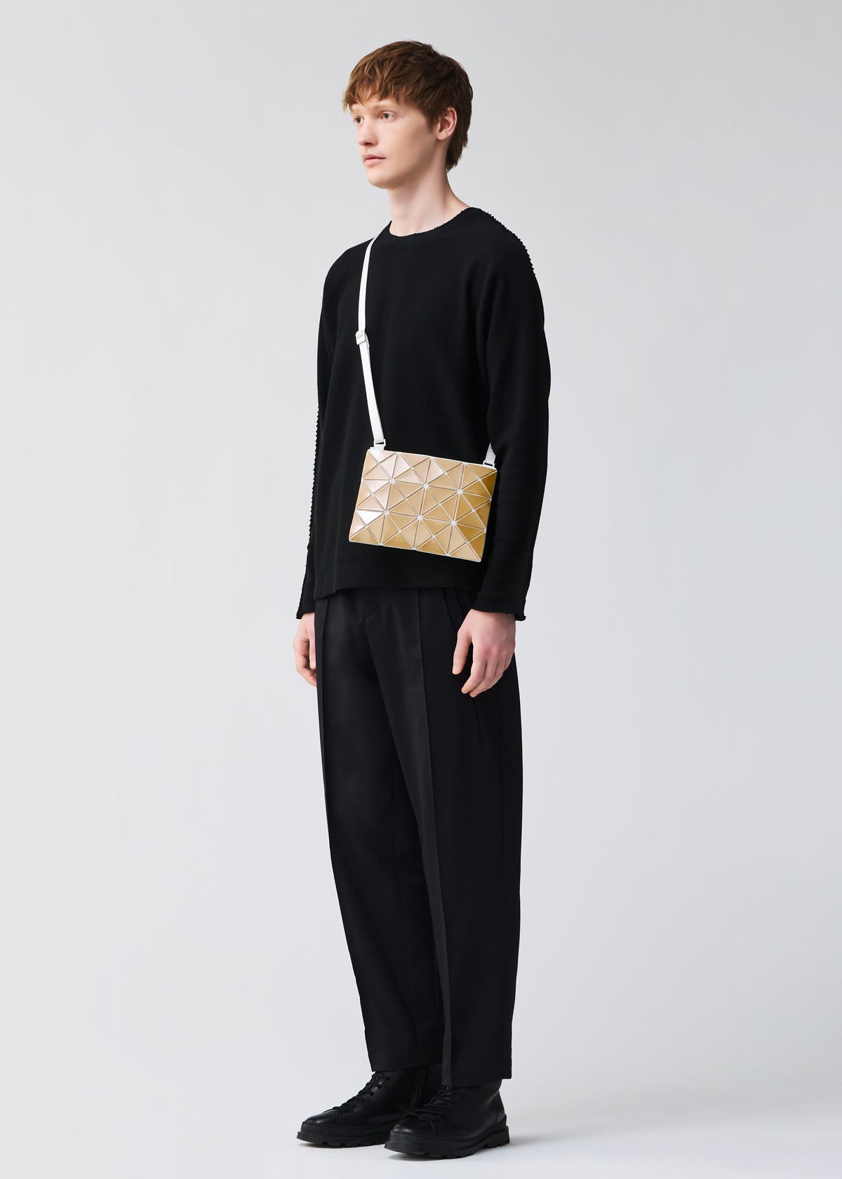 A person with short brown hair wears all black and carries the BAO BAO ISSEY MIYAKE LUCENT METALLIC, a gold geometric-patterned shoulder bag with a metallic finish and white strap, against a plain light background. view 10 of 10