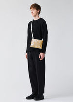 A person with short brown hair wears all black and carries the BAO BAO ISSEY MIYAKE LUCENT METALLIC, a gold geometric-patterned shoulder bag with a metallic finish and white strap, against a plain light background.