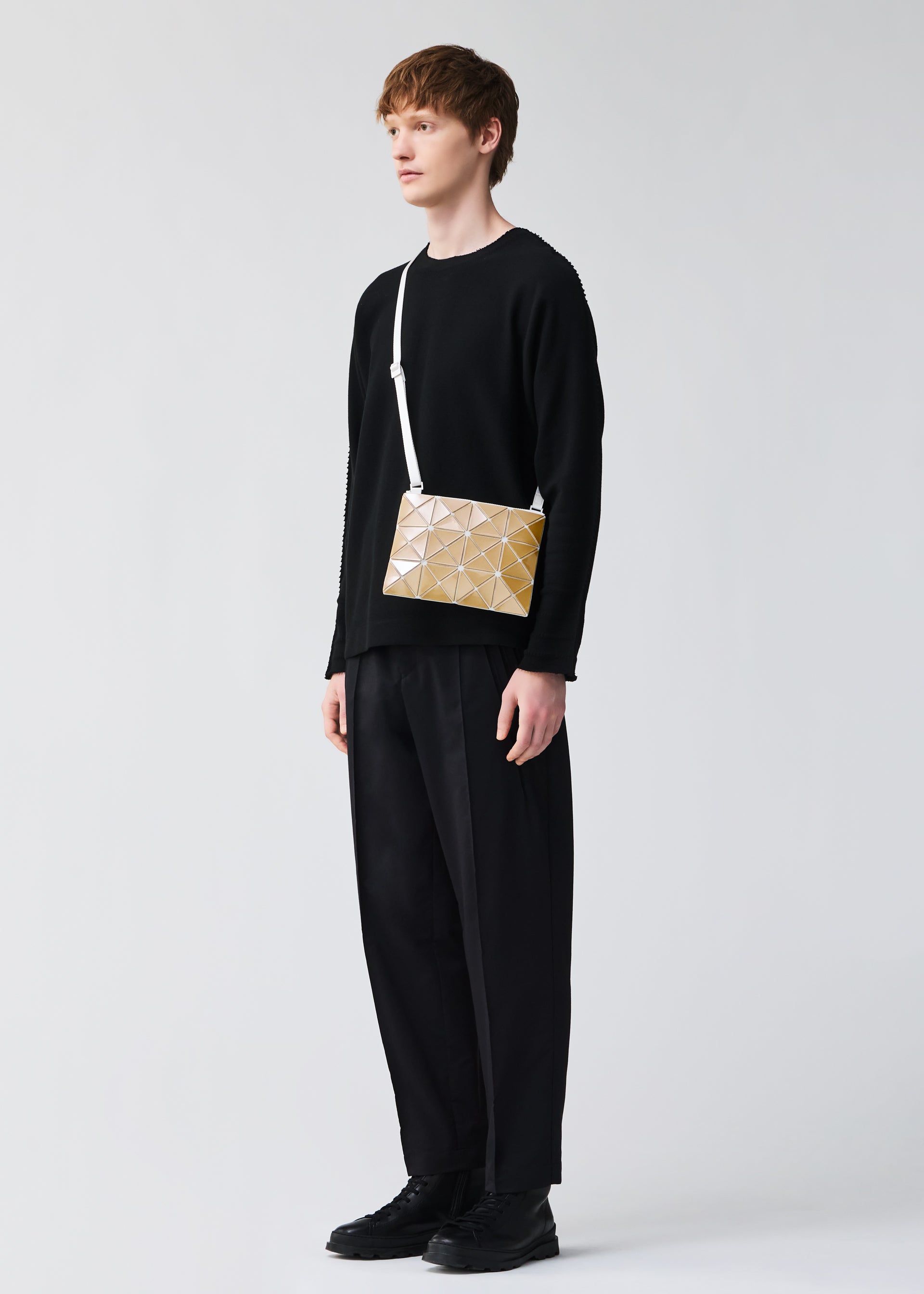 Zoomed view A person with short brown hair wears all black and carries the BAO BAO ISSEY MIYAKE LUCENT METALLIC, a gold geometric-patterned shoulder bag with a metallic finish and white strap, against a plain light background.